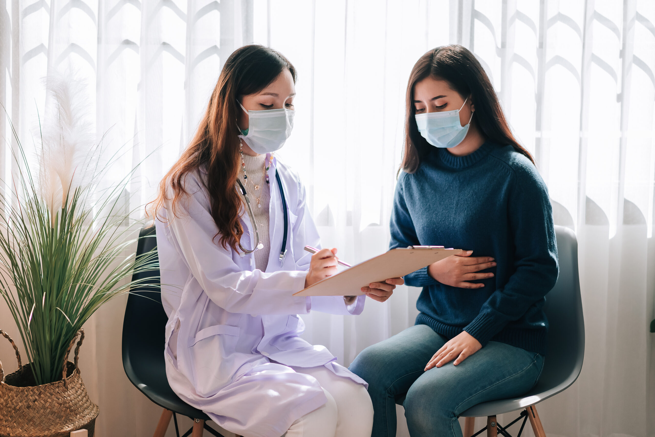 asian doctor visit examines young adult woman hospital with stomach ache doctor checking up consulting health care wearing mask protect covid19 pandemic scaled
